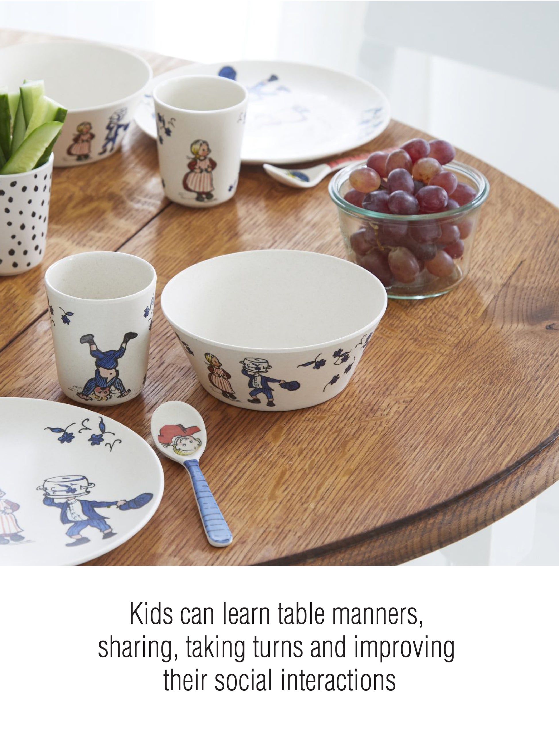Children's tableware set with classic character illustrations, including cups, bowls, plates, and a spoon, arranged on a wooden table with grapes and cucumber sticks. Text on image: 'Kids can learn table manners, sharing, taking turns and improving their social interactions'.