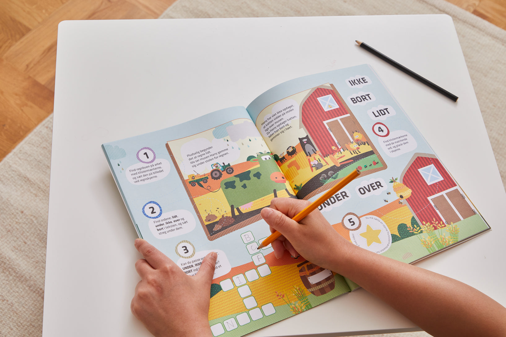 A child is working on an interactive activity book with a farm-themed design on a white table. The book contains colorful illustrations of a barn, animals, and a tractor, along with text-based exercises and word puzzles. The child is holding a yellow pencil and filling in a crossword-style activity, with another black pencil resting on the table. The book includes Danish words such as 'IKKE,' 'BORT,' and 'LIDT,' indicating a language-learning or educational purpose. The background features a light wooden fl
