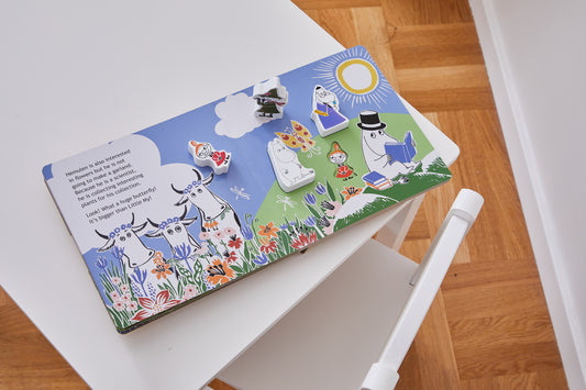 A colorful children's book featuring Moomin characters is open on a white wooden chair with a modern design. The book has interactive wooden pieces depicting various Moomin characters, including Little My, Moomintroll, and Snufkin, placed on a vibrant illustration of a meadow with flowers, butterflies, and a bright sun. The left page contains a short story about Hemulen’s interest in flowers and collecting plants. The floor beneath the chair is made of light wood, complementing the bright and playful atmosp